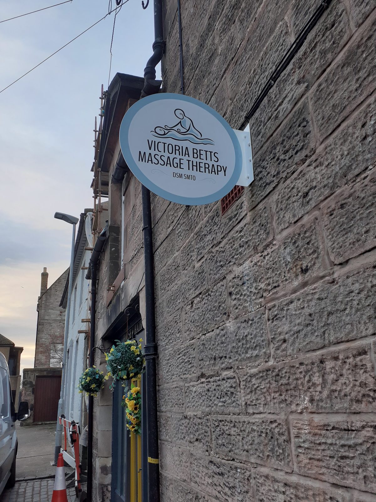 Victoria Betts Massage Therapy's oval hanging sign on the stone wall of Tweed Street