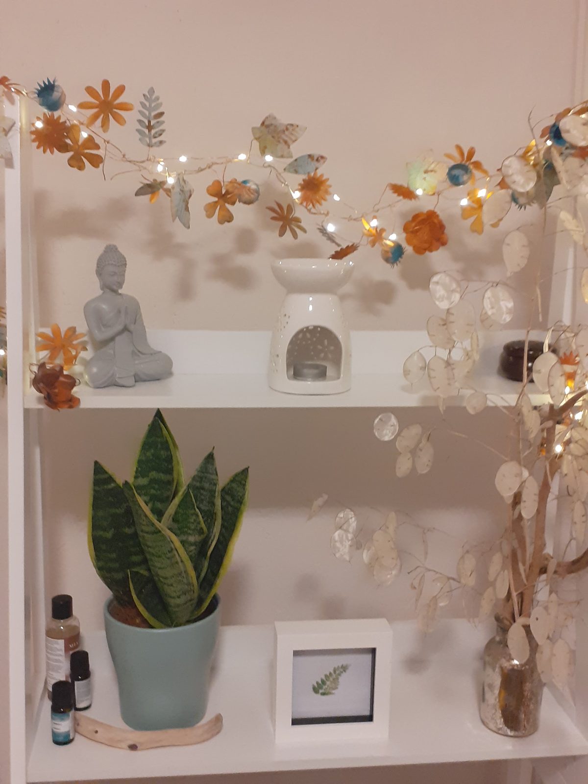 A shelf in the treatment room with a small Buddha, an oil burner, a snake plant, oil bottles and a paper-flower garland with fairy lights